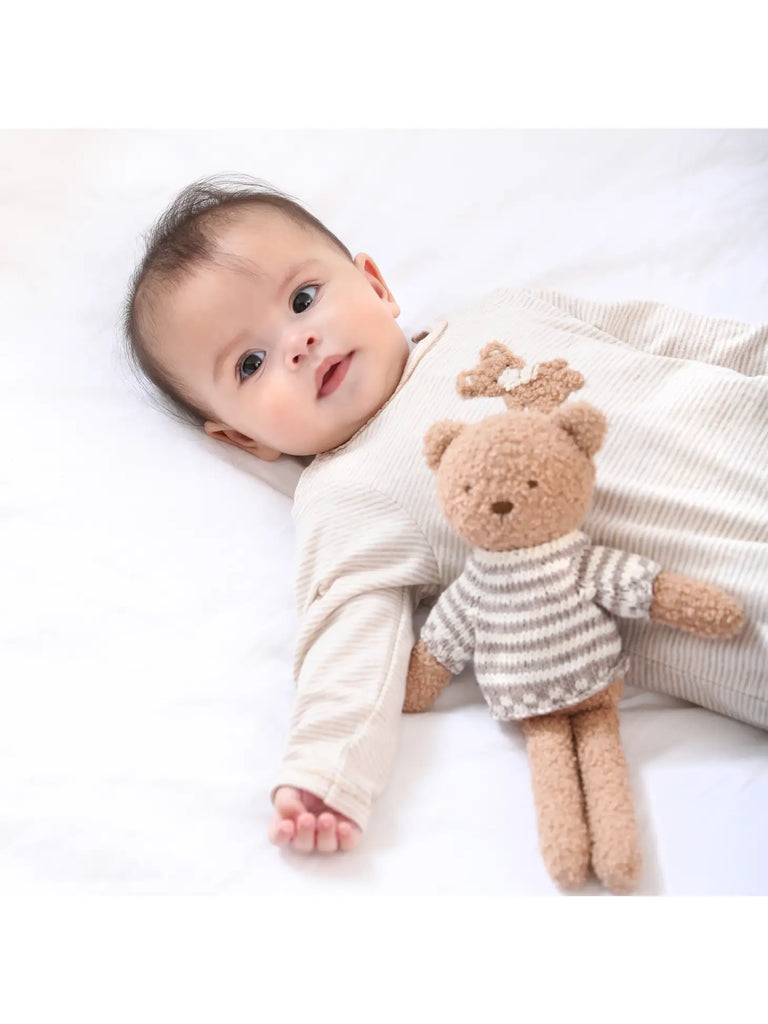 A baby lying on a white blanket, holding a Teddy Bear, looking directly at the camera with a gentle expression. The baby is wearing a striped beige outfit.