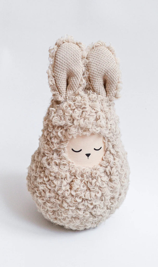 A handmade musical roly poly - Bunny shaped like a sleepy bunny with long ears, covered in soft, curly beige fur, against a plain white background. Its face features closed eyes with a peaceful expression and emits gentle musical sounds.