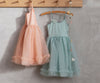 Two Maileg Princess Tulle Dresses - Melon hang on hooks against a light gray wall, like treasures from a magical wardrobe. The dress on the left is peach-colored with ruffled trim, while the dress on the right is light blue-green with similar ruffled detailing. Both are suspended on wooden hangers.