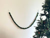 A 6' Felt Holly Garland adorned with small red ornaments drapes gracefully on a white wall. To the right, a section of a Christmas tree with green branches and pinecones is visible, enhancing the enchanting Christmas decor.