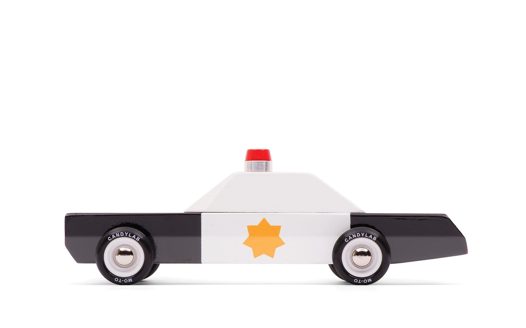 The Candylab Police Cruiser, a minimalist toy police car made of solid beech wood, is showcased on a white background. The vehicle features a black and white color scheme with a yellow star emblem on the door and a small red light on the roof. Its black and silver wheels are equipped with safety-tested rubber tires, adding to its simple and clean design.