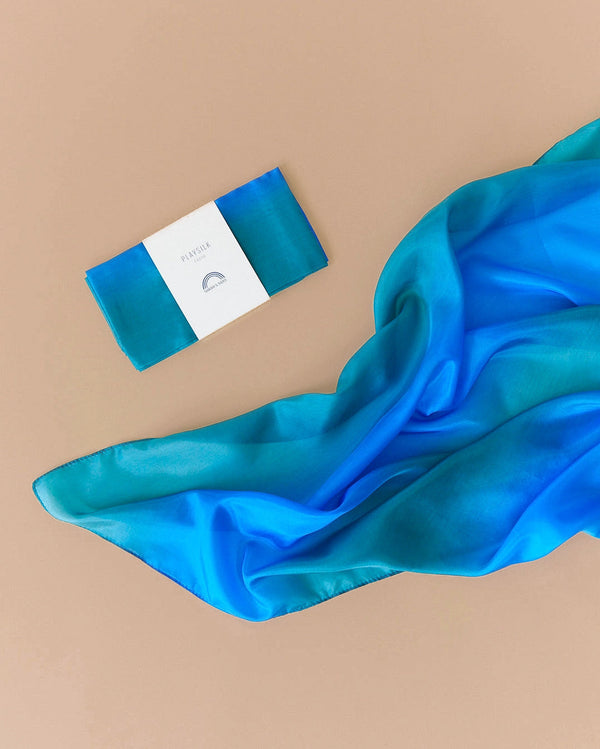 An elegantly draped silk scarf in shades of vibrant blue and turquoise graces a beige surface, while the beautifully packaged Sarah's Silk Enchanted Playsilk - Ocean invites imaginative play with its natural materials.