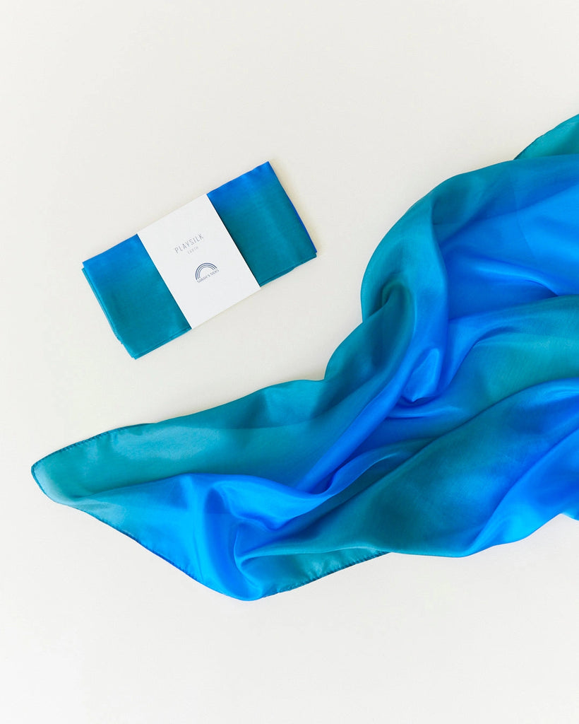 The Sarah's Silk Enchanted Playsilk - Ocean is beautifully showcased against a white backdrop, its vibrant blue and green hues suggesting imaginative play as it flows freely. Beside it, a neatly folded and packaged version with a visible label adds an elegant touch.