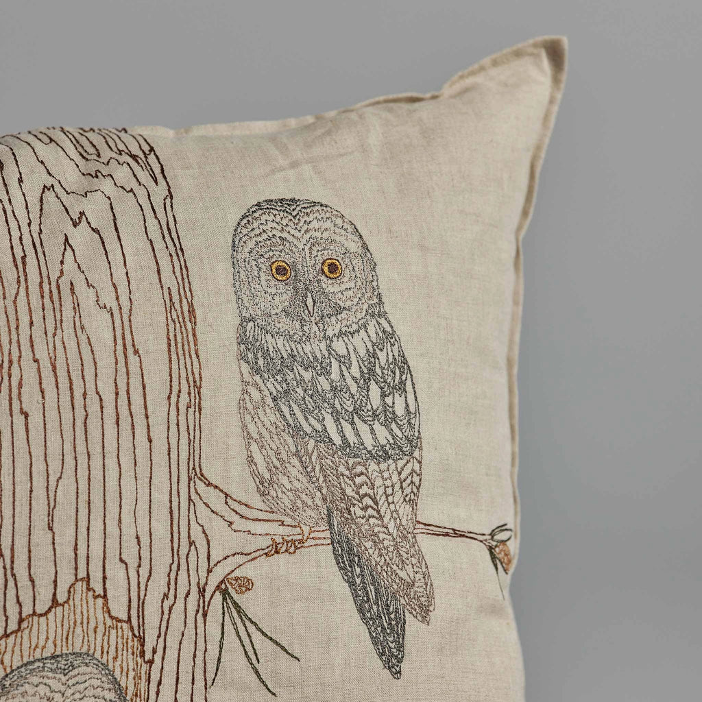 The Coral & Tusk Owl Family Tree Pillow showcases an embroidered design of an owl perched on a tree branch. The owl features detailed grey and brown feathers with striking yellow eyes, and a portion of the tree is visible on the left side of the 100% linen fabric cushion.