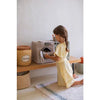 A young girl in a yellow dress is playing with a Basket Honey Pot in a cozy playroom, kneeling on a rug made from recycled PET-bottles.