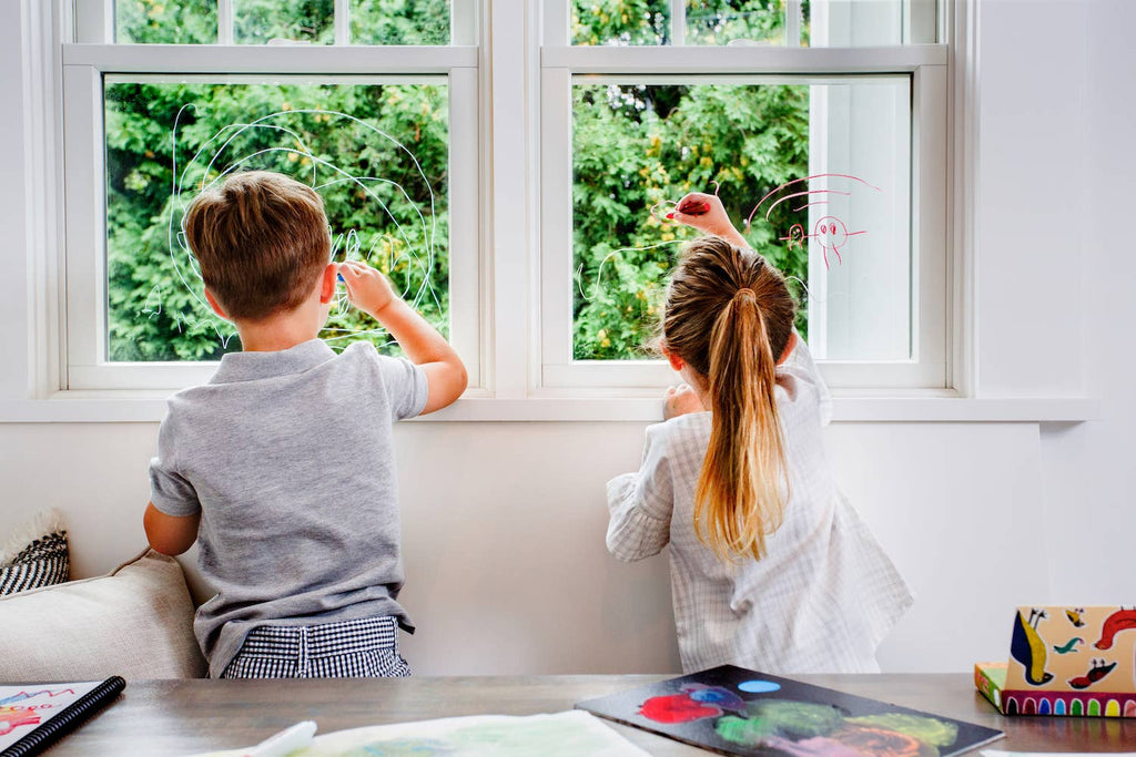 Two children draw on a sunlit window with Kitpas Crayons - 12 Colors, their vibrant artwork framed by the lush green trees outside. Non-toxic art supplies rest on a nearby table, ready for more creative endeavors.