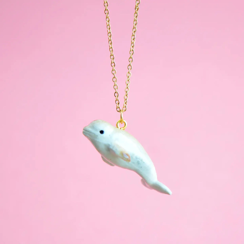 A gold chain necklace is shown against a pink background. The pendant is a small, handcrafted porcelain whale with a subtle glossy finish and a tiny black eye. The necklace appears to be suspended, creating an oceanic elegance for this whimsical White Beluga Necklace.