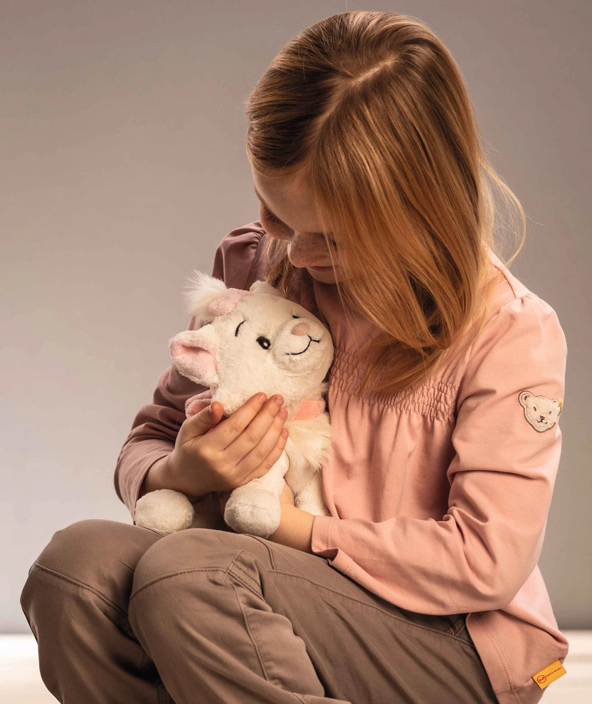 A young girl with light brown hair, wearing a long-sleeved pink shirt and beige pants, is sitting and hugging a Steiff, Disney's "Aristocats" Marie Cat Plush Stuffed Toy, 9 inches. She is looking down at the toy with a gentle and affectionate expression. The background is neutral and softly lit.