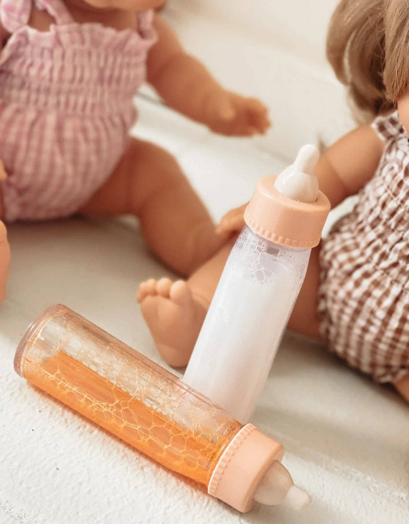 Two Tiny Harlow baby dolls sit on a white surface. In front of them is a Bottled milk and juice set, with one bottle filled with an orange liquid and the other with a milk-like liquid. The dolls are dressed in patterned outfits, one in pink gingham and the other in brown gingham.