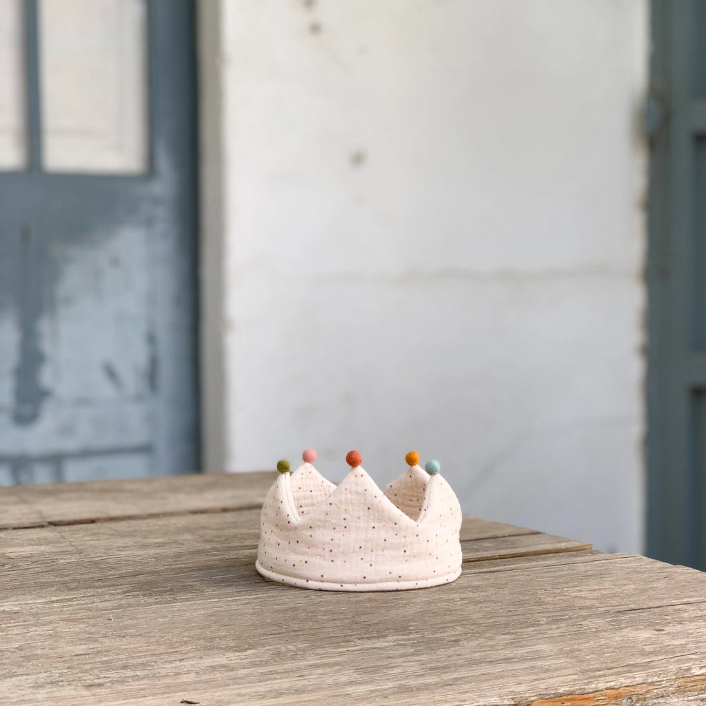 A soft, white handmade Confetti Reversible Birthday Crown made from organic cotton, adorned with colorful beads at the tips of its points, is placed on a rustic wooden table. The background features a worn, light-colored wall and a door with peeling blue paint.