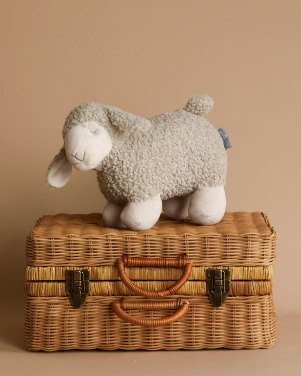 A sleepy Charlotte the Sheep Stuffed Animal snuggles atop a wicker suitcase. Crafted from soft, cuddly fabric, she is perfect for little ones. For easy care, she can be given a gentle machine wash.