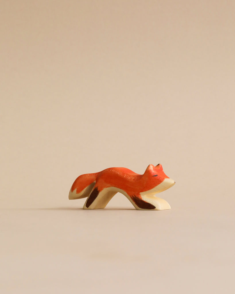 A small, hand-painted Holzwald Small Fox figurine in a leaping pose, placed against a plain, light beige background. The fox is predominantly orange with white details. This high-quality toy