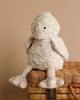 Bill the Duck Stuffed Animal, a fluffy and white plush toy, sits atop a closed wicker basket. This adorable companion features soft wings, a small beak, and large floppy feet, with its charm enhanced by a neutral background.