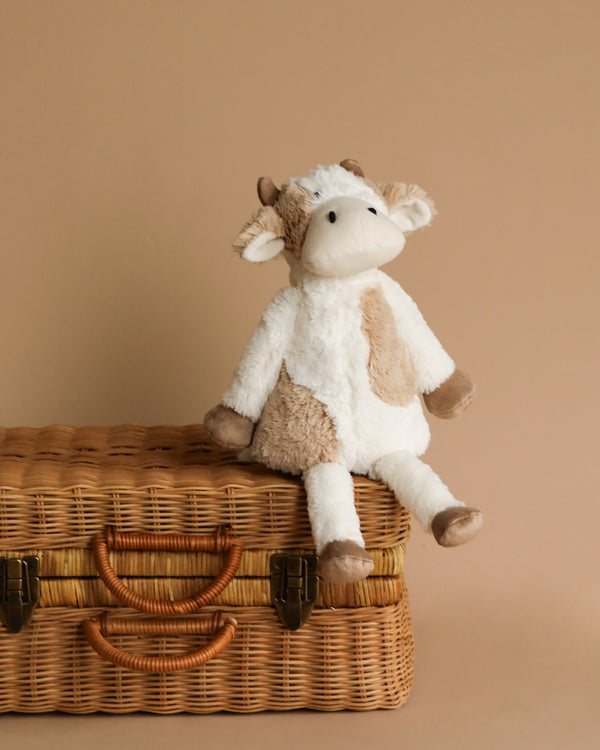 Clover The Cow Stuffed Animal rests atop a wicker suitcase, making for the perfect snuggly toy or friendship gift.