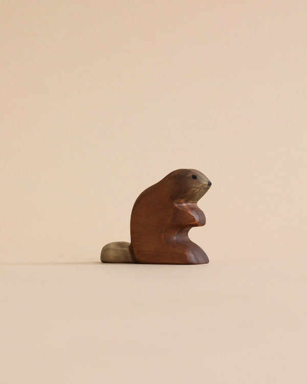 A small, wooden carving of a beaver sits against a plain, light brown background. The beaver is depicted in a sitting position with its front paws folded. Its tail is textured, contrasting slightly with the smooth surface of its body. This piece exemplifies the charm of the Handmade Holzwald Beaver.