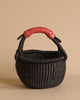 The Handmade Explore Basket is a round, dark woven basket made from elephant grass, featuring a sturdy handle wrapped in red material. Hand-made in Bolgatanga, Ghana, it boasts a tightly braided texture and is displayed against a plain beige background.