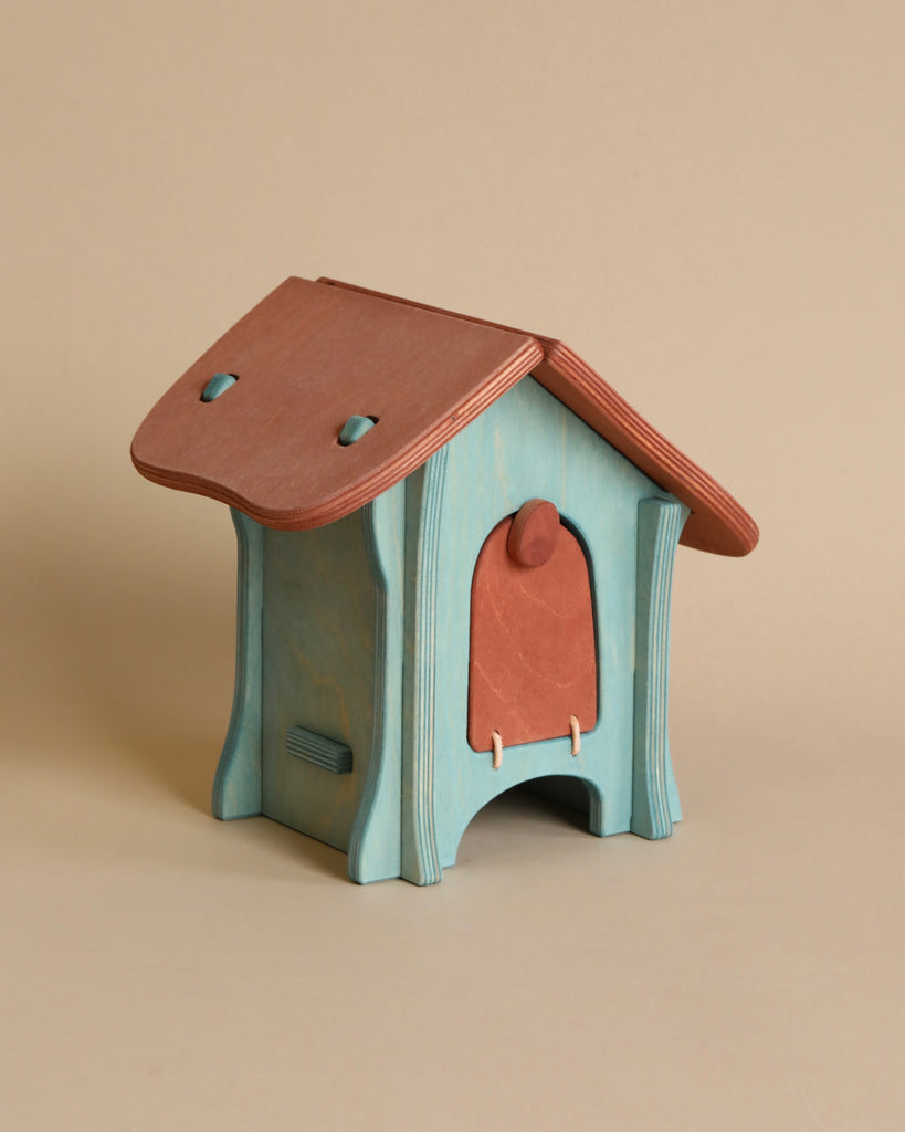 A small, whimsical wooden house with a teal base and a reddish-brown roof, reminiscent of an Ostheimer Hen House / Chicken Coop. The front features a small, arched doorway with a triangular pediment above it. The house has curved edges and a playful design, perfect for imaginative play, all set against a neutral beige background.