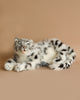 A Snow Leopard Stuffed Animal, known for its unique personality, is lying down on a light brown surface, looking directly at the camera with its tongue sticking out. This charming toy has grey and white fur with black spots and stripes.