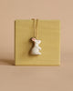 The White Rabbit Necklace features a 24k gold plated pendant shaped like a small white rabbit with pink ears, showcased against a light yellowish-brown square background. The minimalistic design and arrangement highlight the heirloom quality of the piece, set against a plain beige backdrop.