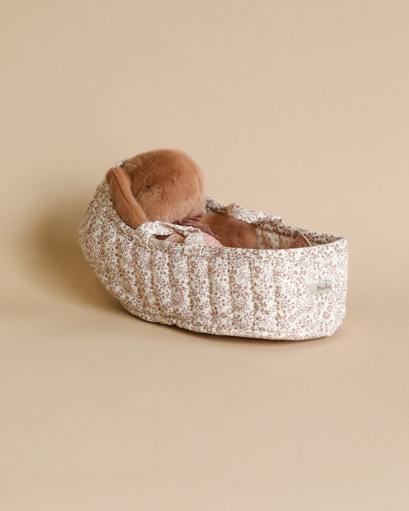 A cozy Maileg Carry Cot, Large in off white, adorned with a floral pattern and featuring a warm knitted blanket alongside a fuzzy brown pillow, set against a neutral beige background.