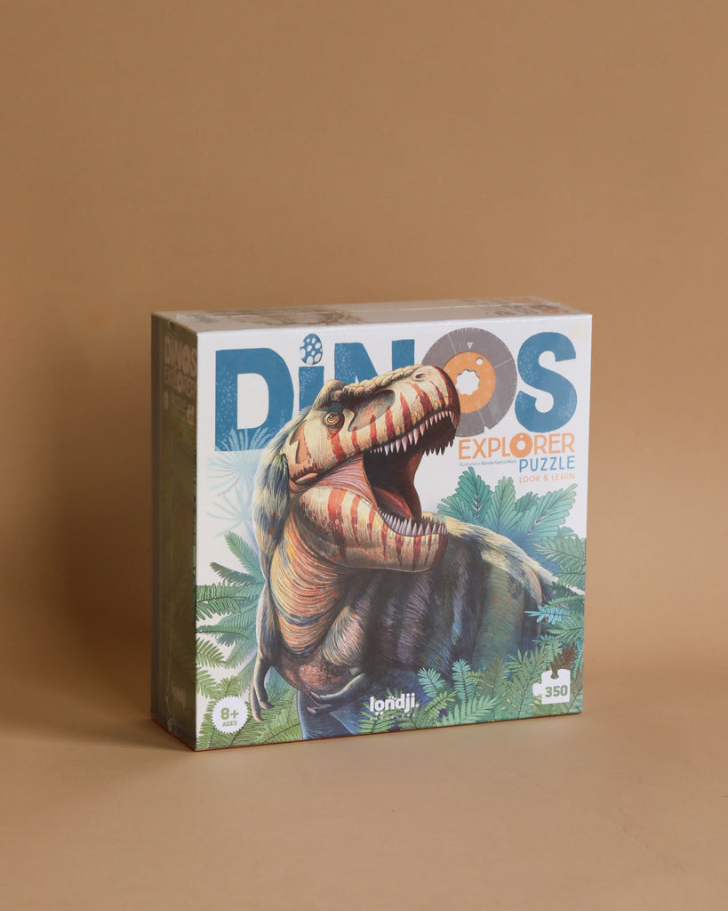 A box of the "Londji - Dino Explorer Puzzle (350 pcs)" is displayed against a beige background. The box showcases an illustration of a roaring dinosaur surrounded by foliage. This 350-piece, educational dinosaur puzzle is designed for ages 8 and up, making it the perfect sustainable toy for young explorers.