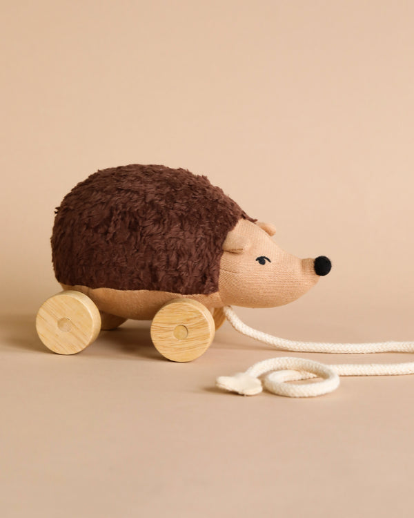 Meet the Hedgehog Pull Toy, a plush toy with a textured dark brown body and light brown face, made from organic cotton. Mounted on four wooden wheels, she features a white pull rope with a loop handle. Set against a solid beige background, this hedgehog adds charm to any enchanted forest adventure.