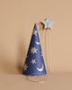 A blue, sparkly Meri Meri Wizard party hat adorned with silver stars and crescent moons stands upright on a beige surface. Beside it is a matching magic wand featuring a silver star on a wooden stick. A thin elastic strap at the base of the hat indicates where it can be worn.