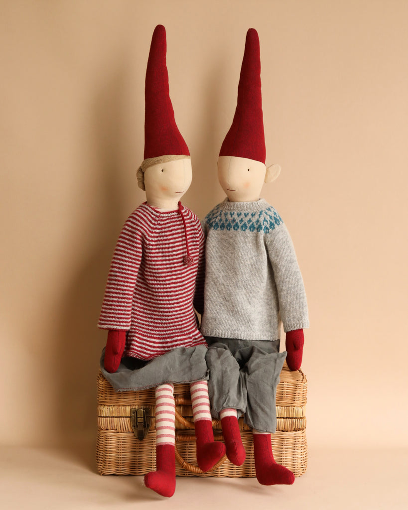 Two decorative Maileg Christmas Mega Pixy dolls seated on a wicker basket, one wearing a striped red and white shirt and the other in a grey and blue sweater, both with long red hats.