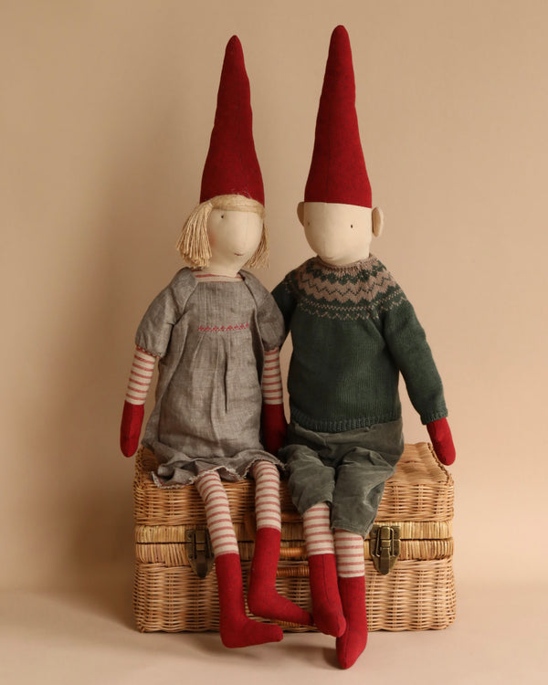 Two Maileg Christmas Pixy (Size 5) dolls with pointed red hats sitting side by side on a wicker basket, against a neutral background. The dolls feature striped leggings, intricate clothing made from exclusive materials, and stitched