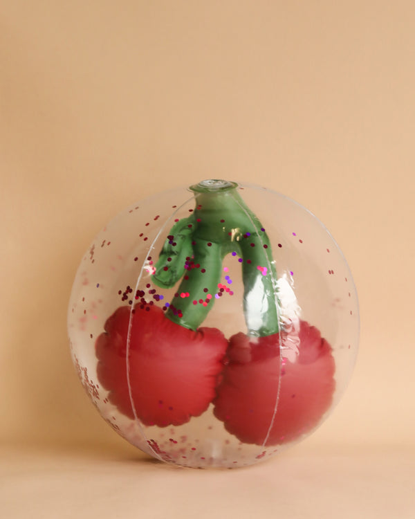 A glass paperweight with a realistic Inflatable Cherry Beach Ball design inside, featuring vibrant red color and green detailing, set against a plain beige background.