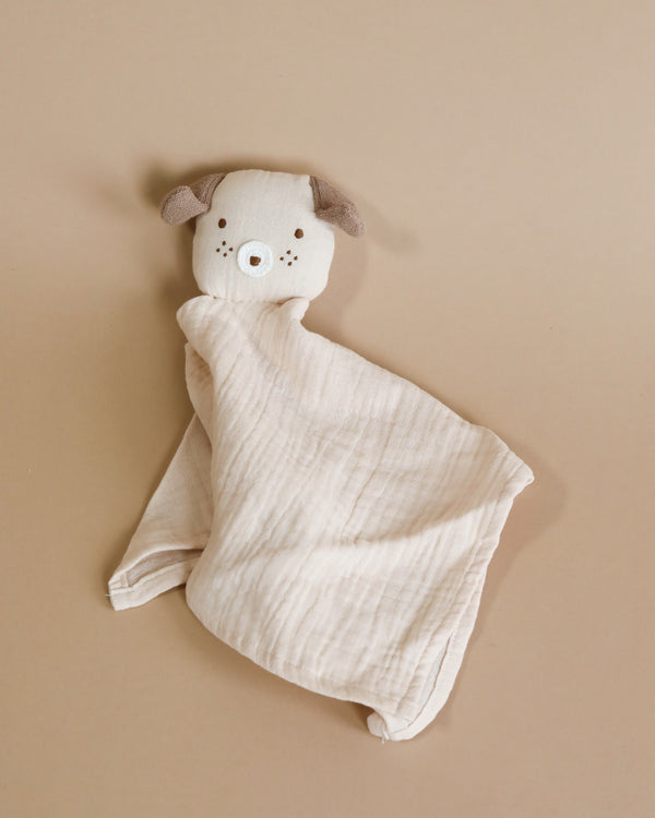 Introducing the Dog Lovey: a baby security blanket featuring a cute, plush dog's head. The dog has light brown ears, small embroidered eyes, and a nose. The blanket body is made from light beige textured organic cotton. The background showcases a smooth, neutral brown surface.