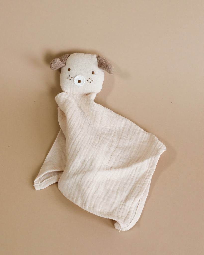 Introducing the Dog Lovey: a baby security blanket featuring a cute, plush dog's head. The dog has light brown ears, small embroidered eyes, and a nose. The blanket body is made from light beige textured organic cotton. The background showcases a smooth, neutral brown surface.