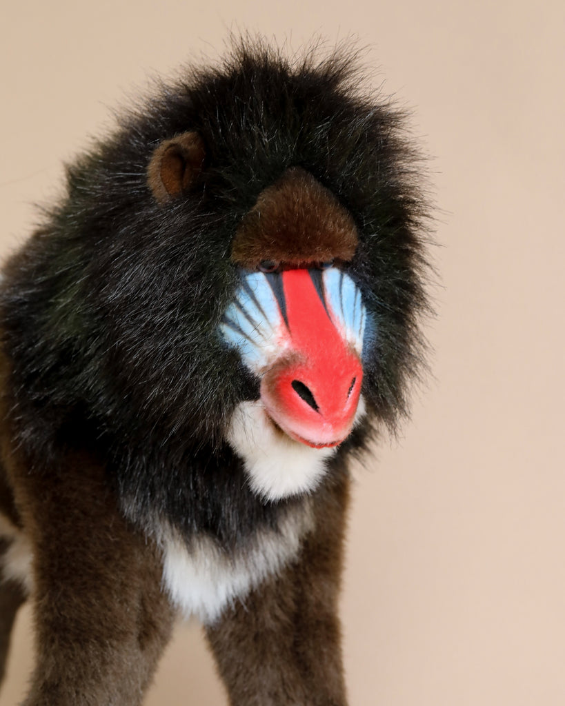 A close-up of the Hansa Mandrill Monkey Stuffed Animal, featuring a hand-sewn design with a vibrant red and blue snout and dark fur, showcasing realistic features, set against a plain beige background.