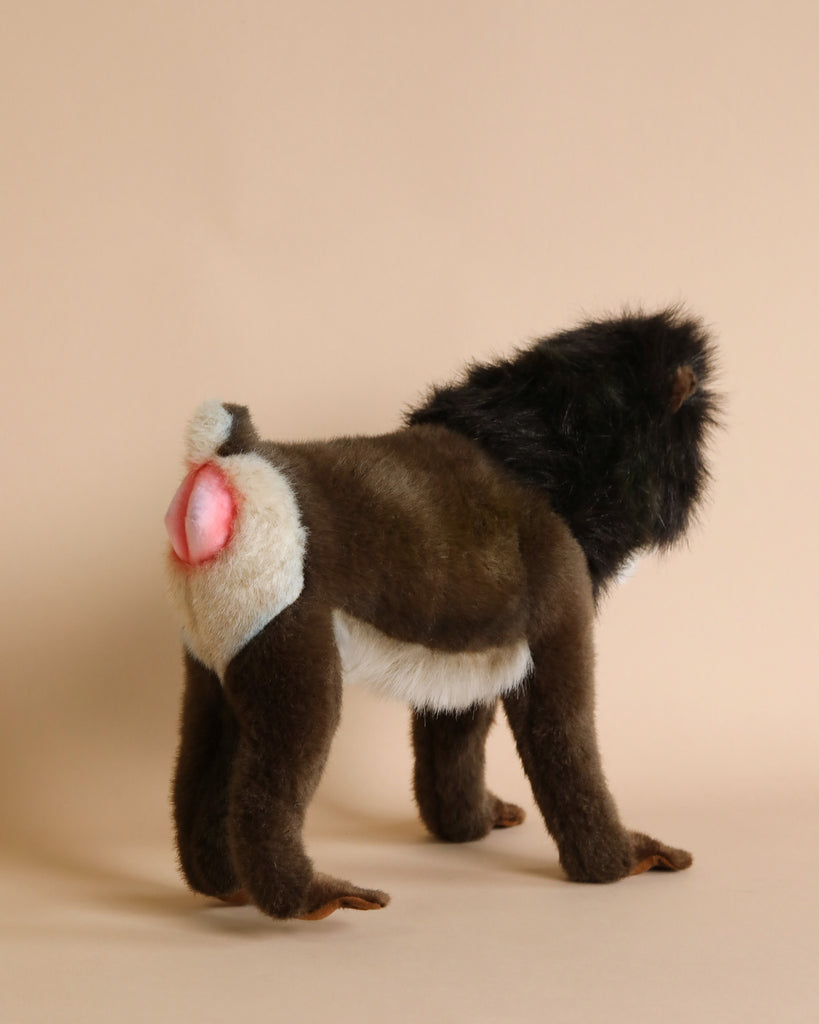 A lifelike model of a Hansa Mandrill Monkey Stuffed Animal stands on all fours against a plain beige background. The mandrill, hand sewn with realistic features, has dark brown fur, a black mane around its head, and prominent pink and white coloring on its hindquarters.