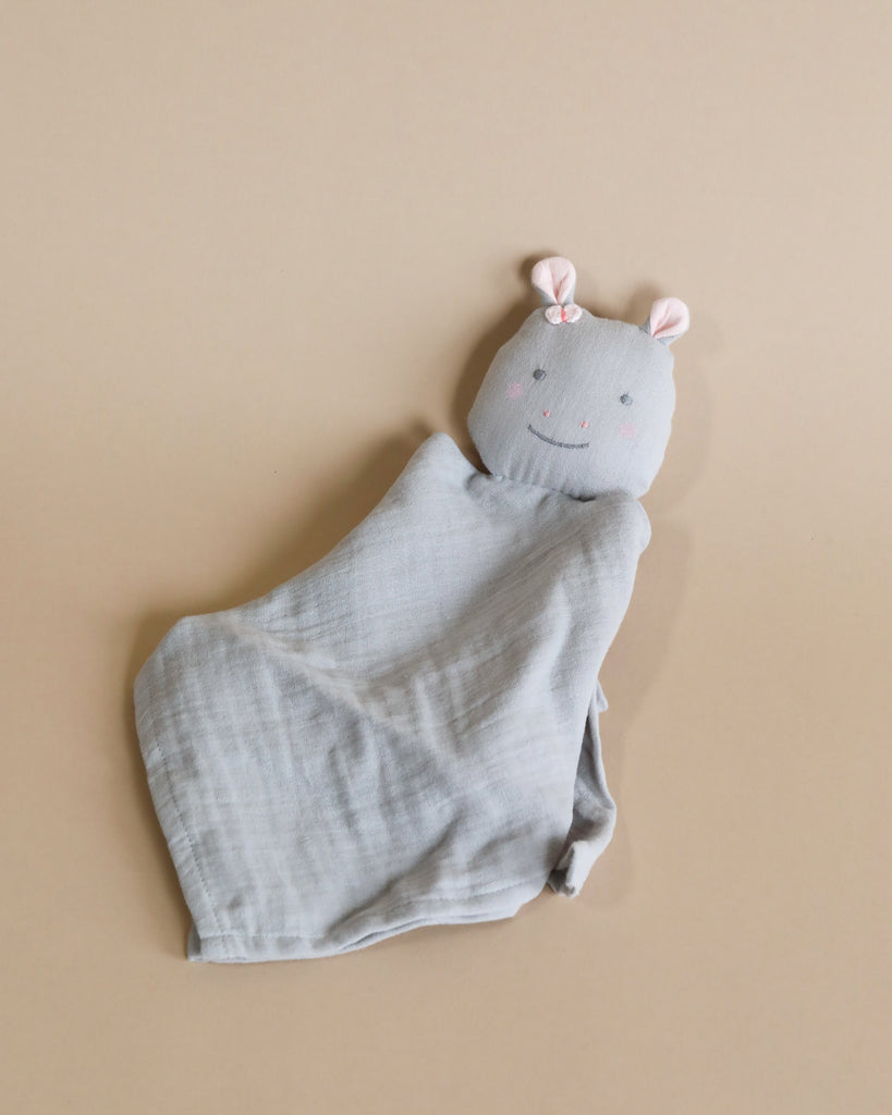 A cute, soft gray Hippo Lovey made from organic cotton with a plush animal head at one end. The plush head has a smiling face, pink blush cheeks, and small pink ears. Filled with polyester filling for extra softness, the blanket is laid out on a beige surface.