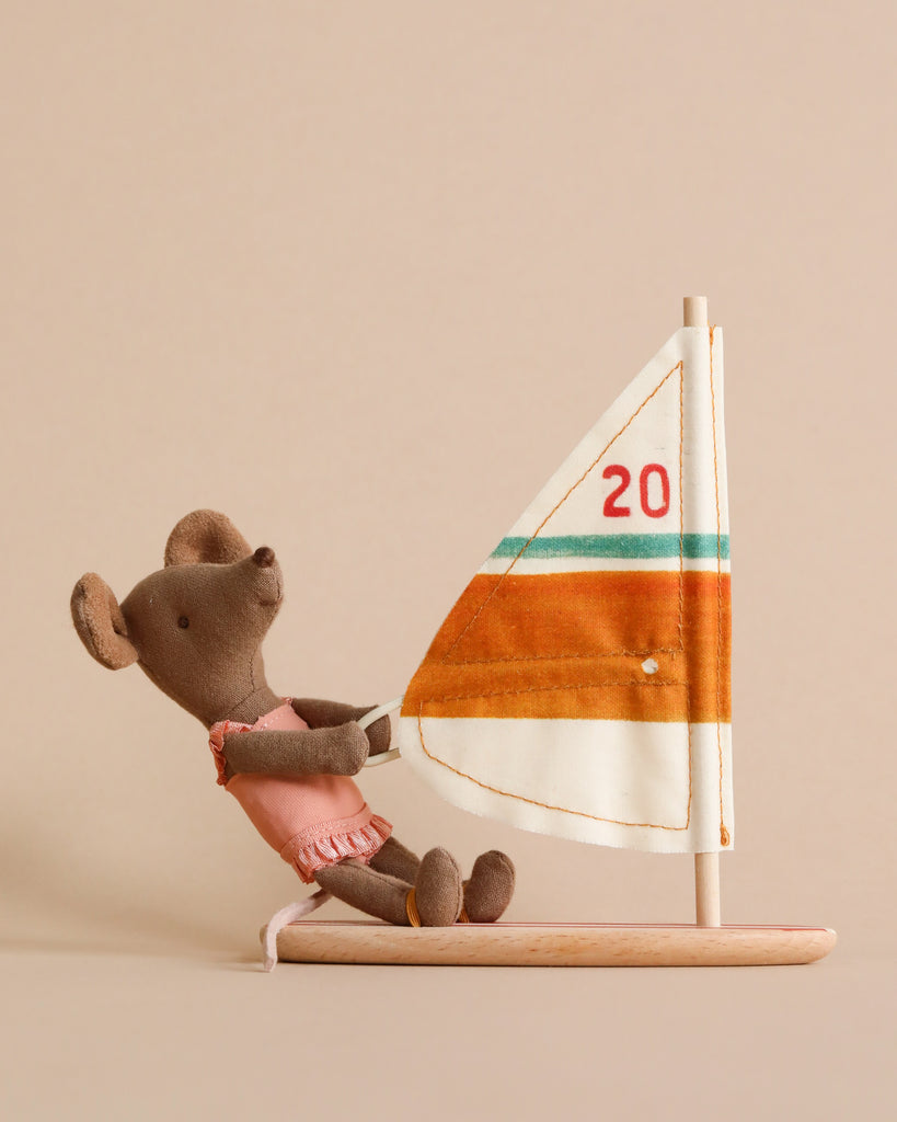 A Maileg Little Sister Surfer Mouse wearing a pink outfit is sitting on a wooden platform, holding onto an orange, white, and brown striped sail with the number 20 on it. This adorable surfing toy features magnetic hands for better grip. The background is beige.