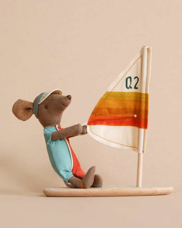 A Maileg Surfer Big Brother Mouse in a blue and red outfit with a cap sits on a wooden surfboard featuring the text "Q2" and orange and yellow stripes. The mouse, looking like one of those surfer mice, is holding the sail's pole. The background is plain beige.