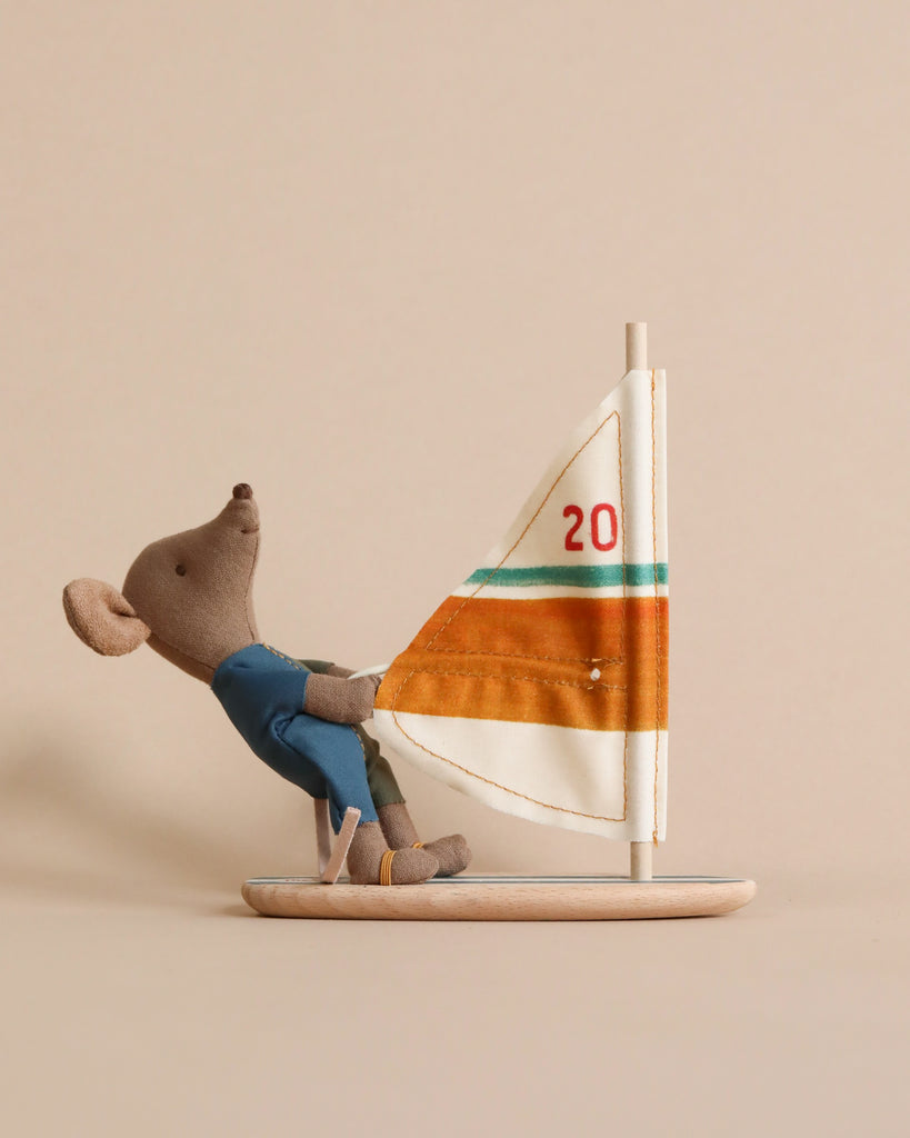 A Maileg Little Brother Surfer Mouse in a blue wetsuit is sitting on a small wooden sailboat with an orange and white sail marked with the number 20. The background is a plain beige color.