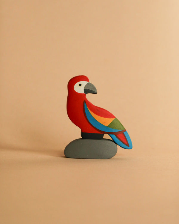 The "Handmade Wooden Parrot On A Rock" features a red body, gray beak, and multicolored wings with a hand-painted finish, perched on a smooth oval stone against a plain beige background.