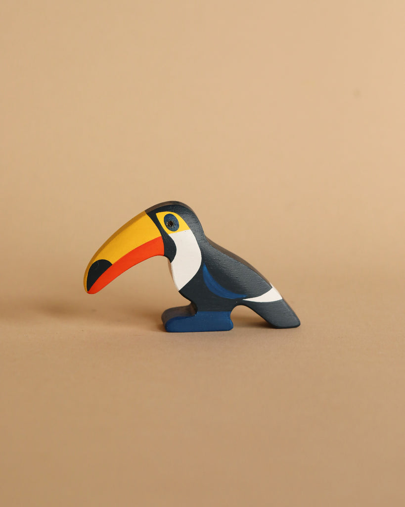 The Handmade Wooden Toucan figurine is crafted with a hand-painted finish, showcasing a prominent yellow beak and vibrant blue, white, and orange patterns on a beige backdrop. It's an ideal addition for small world play and adds charm to any collection.