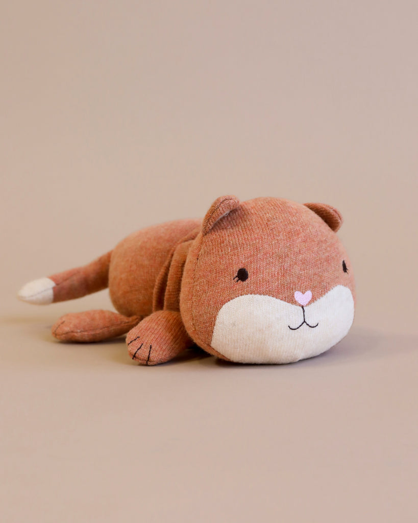 A soft, brown Harmonica Fox Activity Toy made from recycled polyester is lying on its stomach against a neutral background. The toy has a white snout with a small pink heart-shaped nose, black stitched eyes, and a small, friendly smile. Its paws and tail are also brown with white accents.