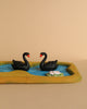 The Handmade Wooden Pond - Black Swans features two petite black wooden swan figurines gracefully floating on a small blue pond, encircled by a golden yellow border. A delicate pink and white lily adorns the pond's edge, set against a neutral beige backdrop that enhances the tranquil ambiance of the scene.