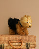 A small Yorkshire Terrier Dog Stuffed Animal stands on a wicker basket with brass clasps, set against a beige background. The hand sewn toy has a fluffy black and tan coat with realistic features and appears to be looking to the side.