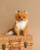 The Sitting Fox Stuffed Animal, a realistic plush toy with reddish-brown fur, a white underbelly, and black-tipped ears, is perched on a wicker basket against a beige background. This unique handmade toy boasts a soft and fluffy appearance, complete with detailed fur and a lifelike expression.