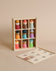 A wooden memory game featuring a grid of eight painted peg dolls in a solid wood box. Each doll is wearing different colorful attire and headgear, encouraging open-ended play. The lid with square cutouts leans against the box, set against a neutral beige background. Perfect for creative play! Introducing the Happy Folk Hotel!