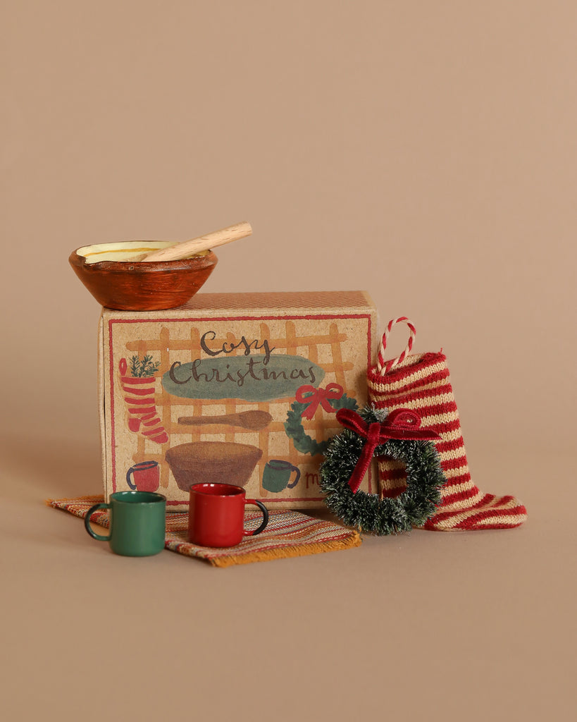 The Maileg Cosy Christmas Set includes a charming array of holiday-themed items such as a decorative box with a painted fireplace, a wooden bowl with a stirrer, a small wreath, festive accessories like a red striped stocking, two small mugs for the mouse family, and a miniature rug all presented on a beige background.