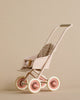 A Maileg Miniature Stroller, Micro - Powder with a pink frame, four cream-colored wheels, and a patterned seat cushion. The stroller is empty and set against a beige background.