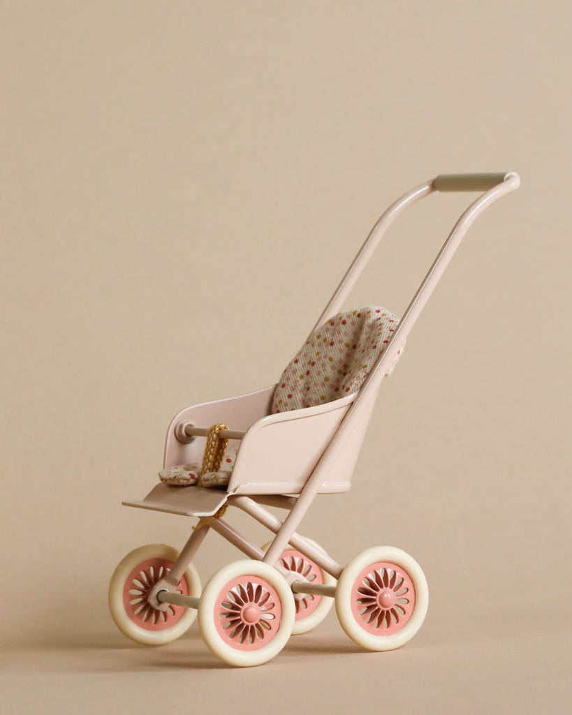 A Maileg Miniature Stroller, Micro - Powder with a pink frame, four cream-colored wheels, and a patterned seat cushion. The stroller is empty and set against a beige background.