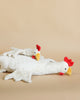 Two Senger Naturwelt Cuddly Animal - Chickens, lying on a beige surface; one stacked on top of the other in a playful manner, both with white bodies and red crests.