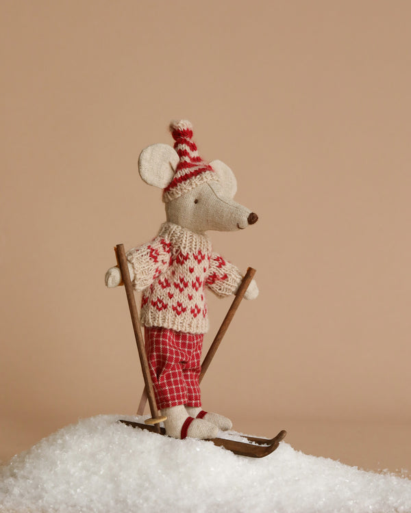 The Maileg Winter Mouse with Ski Set, Mom - Red, features a charming mouse dressed in a red and white sweater, plaid pants, and a striped hat. The mouse balances on two skis with poles atop a small mound of artificial snow set against a plain beige background, perfectly capturing the spirit of snowy adventures.